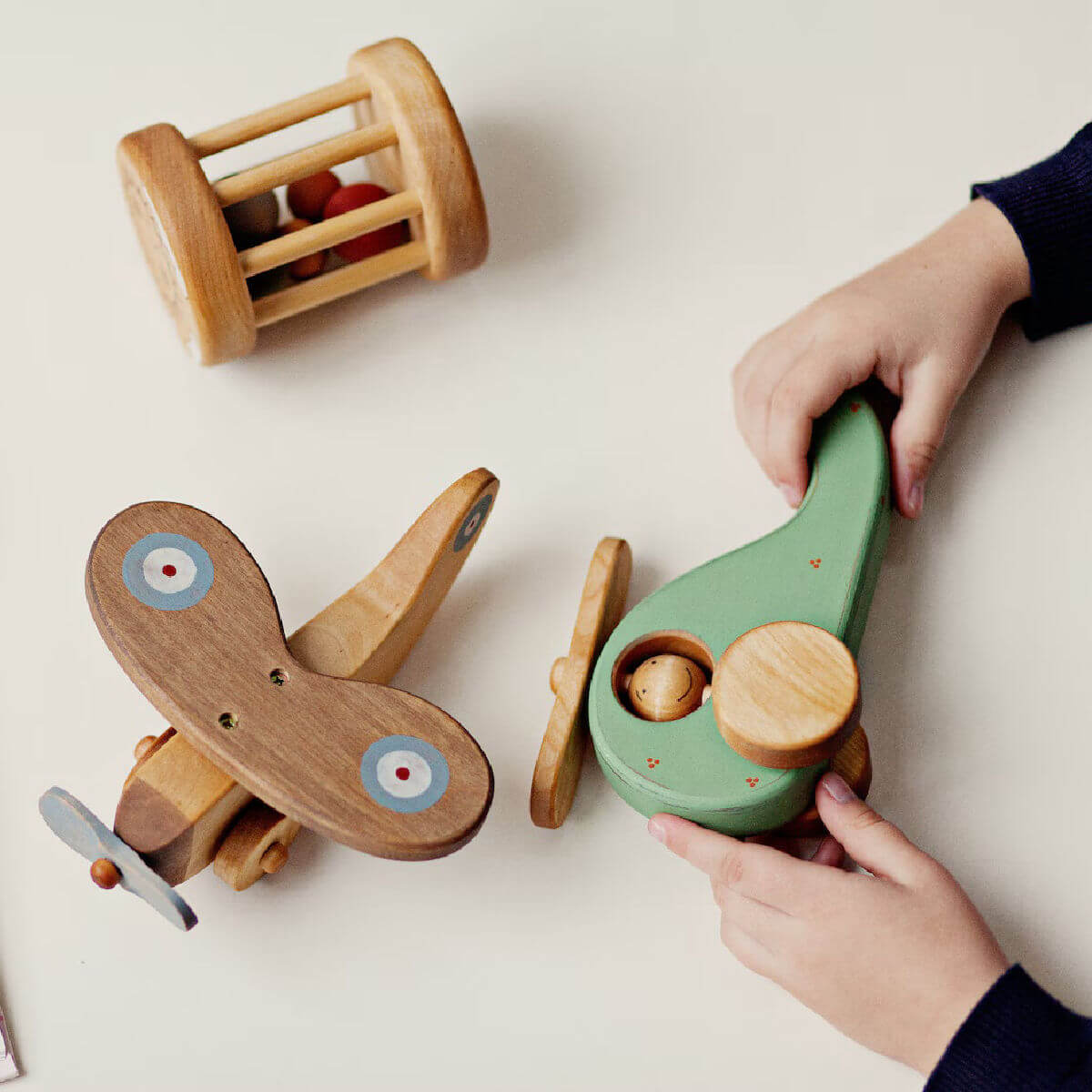 Wooden toys including a plane, car, and green vehicle with a child's hands interacting with them on a light surface.