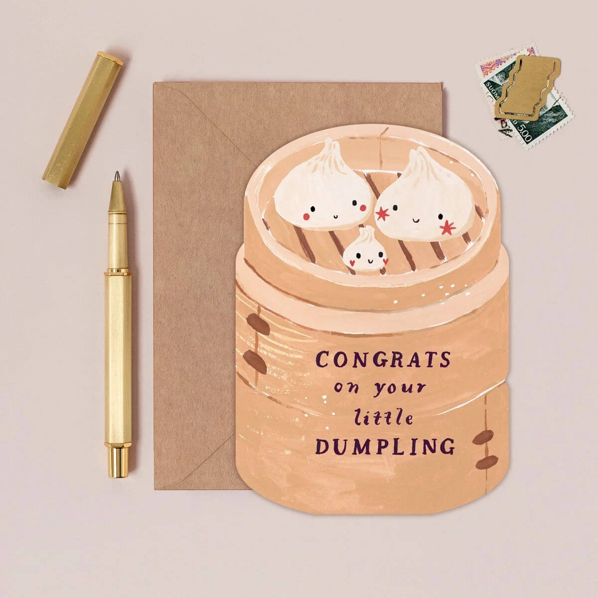 Greeting card with cartoon dumplings in a steamer and 'Congrats on your little dumpling' text, next to a pen and envelope on a beige background.