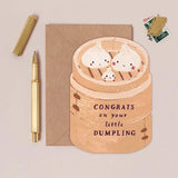 Greeting card with cartoon dumplings in a steamer and 'Congrats on your little dumpling' text, next to a pen and envelope on a beige background.