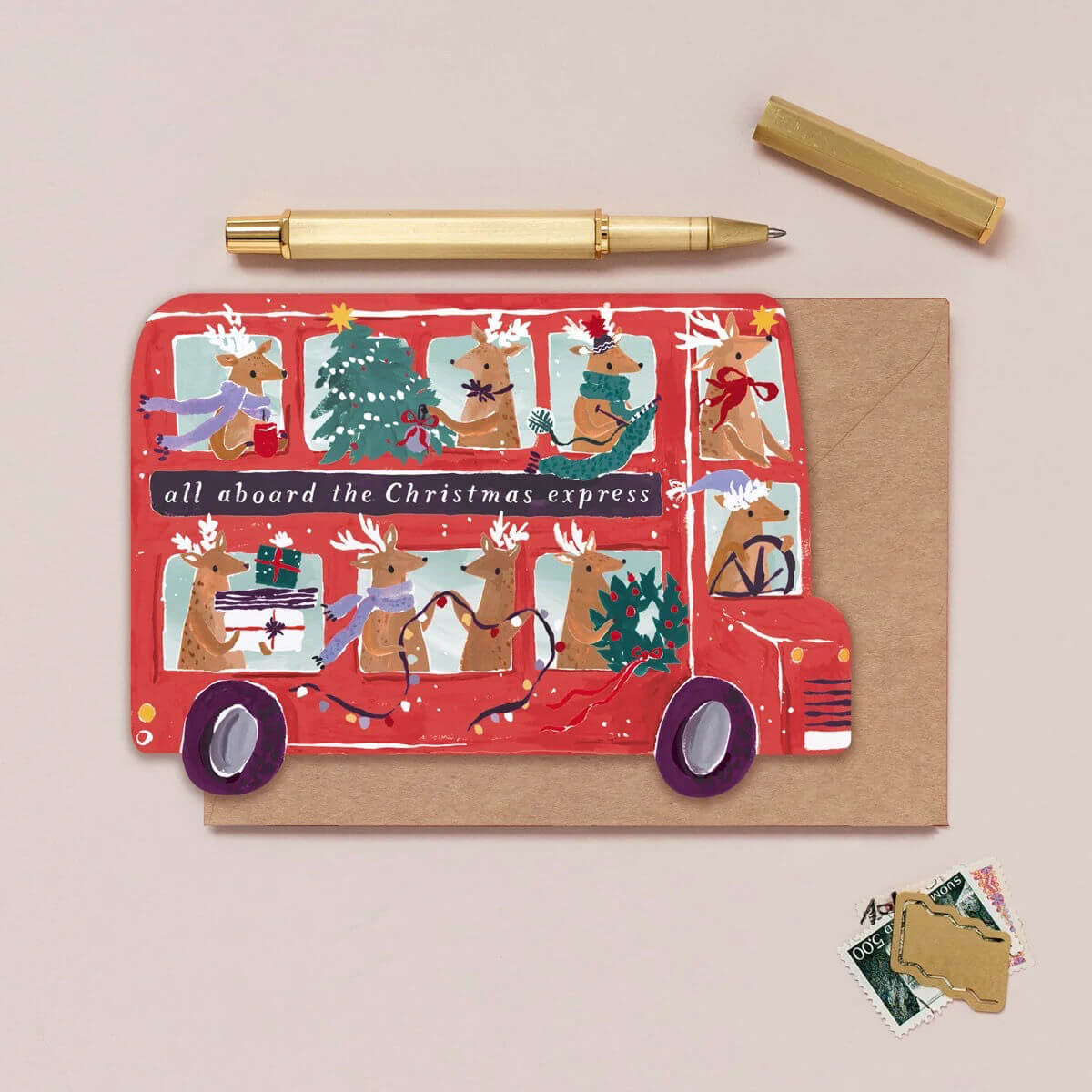 Christmas-themed card with red bus design and reindeer, accompanied by a pen and small envelopes on a beige background.