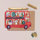 Christmas-themed card with red bus design and reindeer, accompanied by a pen and small envelopes on a beige background.