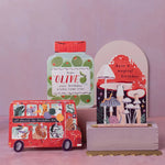 A set of illustrated birthday-themed paper goods and cards on a pink surface against a soft pink background. The set includes a red double-decker bus card, a green bottle-shaped card with the word 'OLIVE', a mushroom card on a stand, and a small round tin with a red lid.