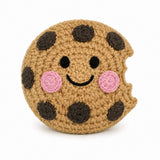 A crocheted plush toy shaped like a chocolate chip cookie with a bite taken out, featuring a smiling face with black eyes and mouth, pink cheeks, and dark brown chocolate chip details on a beige background, set against a plain white studio backdrop.