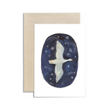 A greeting card with a white front featuring an oval illustration of a white bird flying over a dark blue starry sky, shown with a kraft paper envelope behind it on a white background.