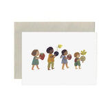 A white greeting card with a minimalist illustration of four children carrying produce, shown with a light beige envelope behind it on a white background.