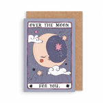 A tarot-style greeting card with a kraft envelope behind it. The card illustration shows a crescent moon and stars with the text 'OVER THE MOON FOR YOU illustrated by sister paper co.