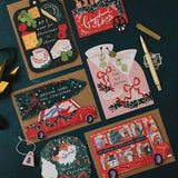 A flat-lay arrangement of Christmas-themed paper cards and a matching ornament on a dark teal background, alongside a gold pen and small decorative items. The cards feature festive illustrations including a red car carrying a Christmas tree, a red double-decker bus, a pink martini glass with holiday garnish, and a black cheese board design.