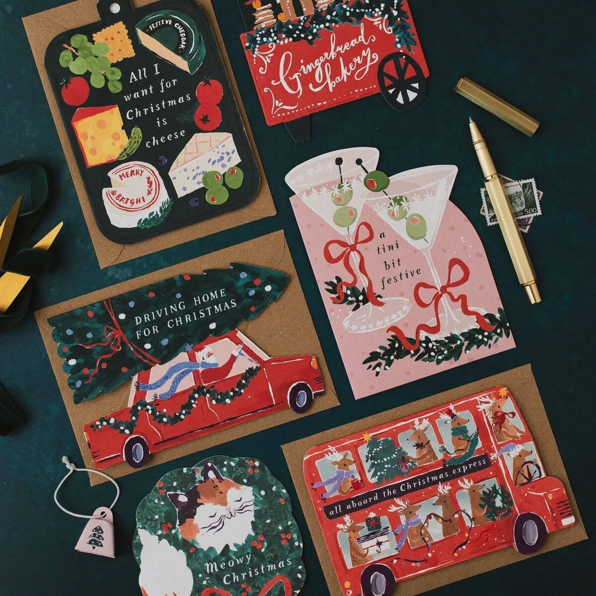 A flat-lay arrangement of Christmas-themed paper cards and a matching ornament on a dark teal background, alongside a gold pen and small decorative items. The cards feature festive illustrations including a red car carrying a Christmas tree, a red double-decker bus, a pink martini glass with holiday garnish, and a black cheese board design.