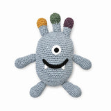 A crocheted monster plush toy in light blue-gray yarn, featuring three colorful antennae in orange, green, and purple, a single large embroidered eye, a black stitched smile, and small white teeth, set against a plain white background.