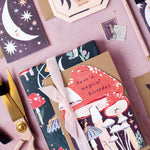 A styled flat-lay of a wrapped gift box on a dusty pink background. The gift is wrapped in dark forest green paper with whimsical mushroom and star illustrations, tied with a pale pink ribbon, and topped with a kraft paper tag that reads 'have a magical birthday' in script over a red mushroom graphic.