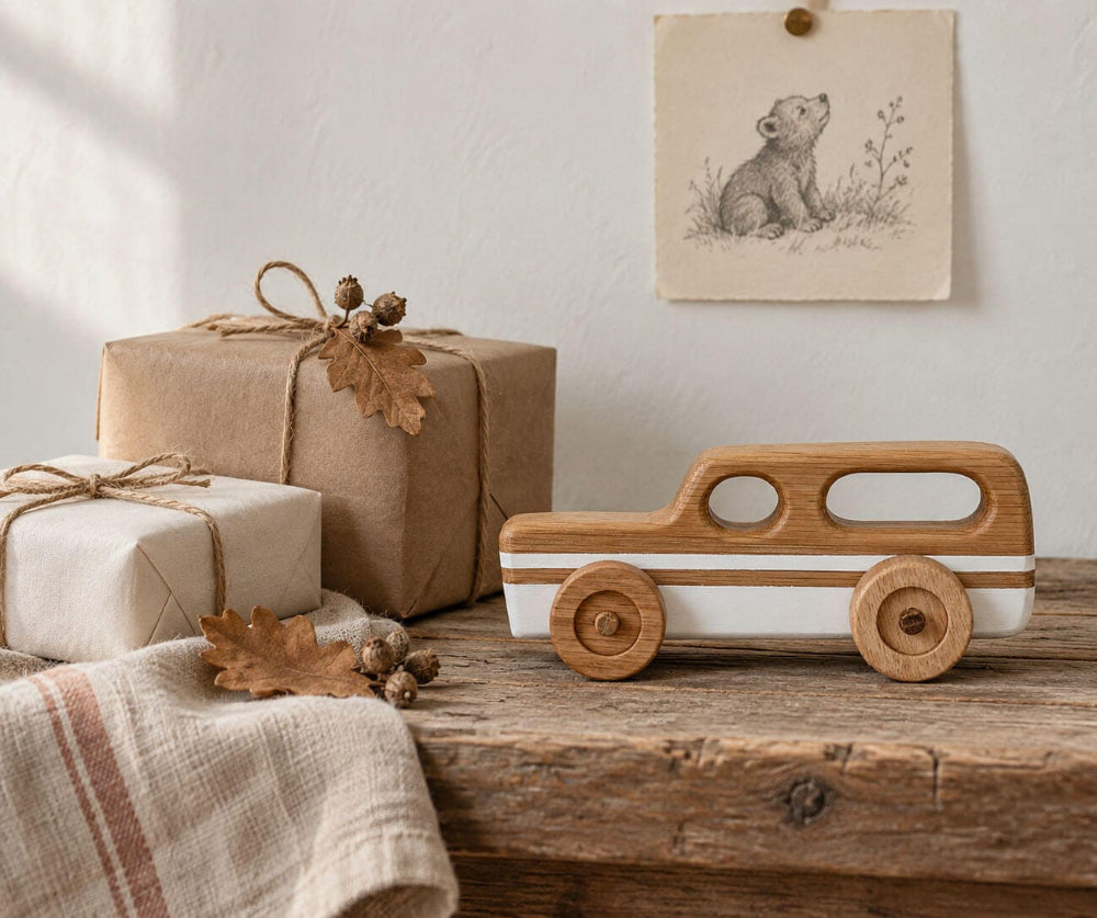 A wooden toy car with a natural oak body and white side stripes sits on a rustic wooden table next to wrapped gift boxes and a linen cloth. A small framed bear illustration hangs on the wall behind.