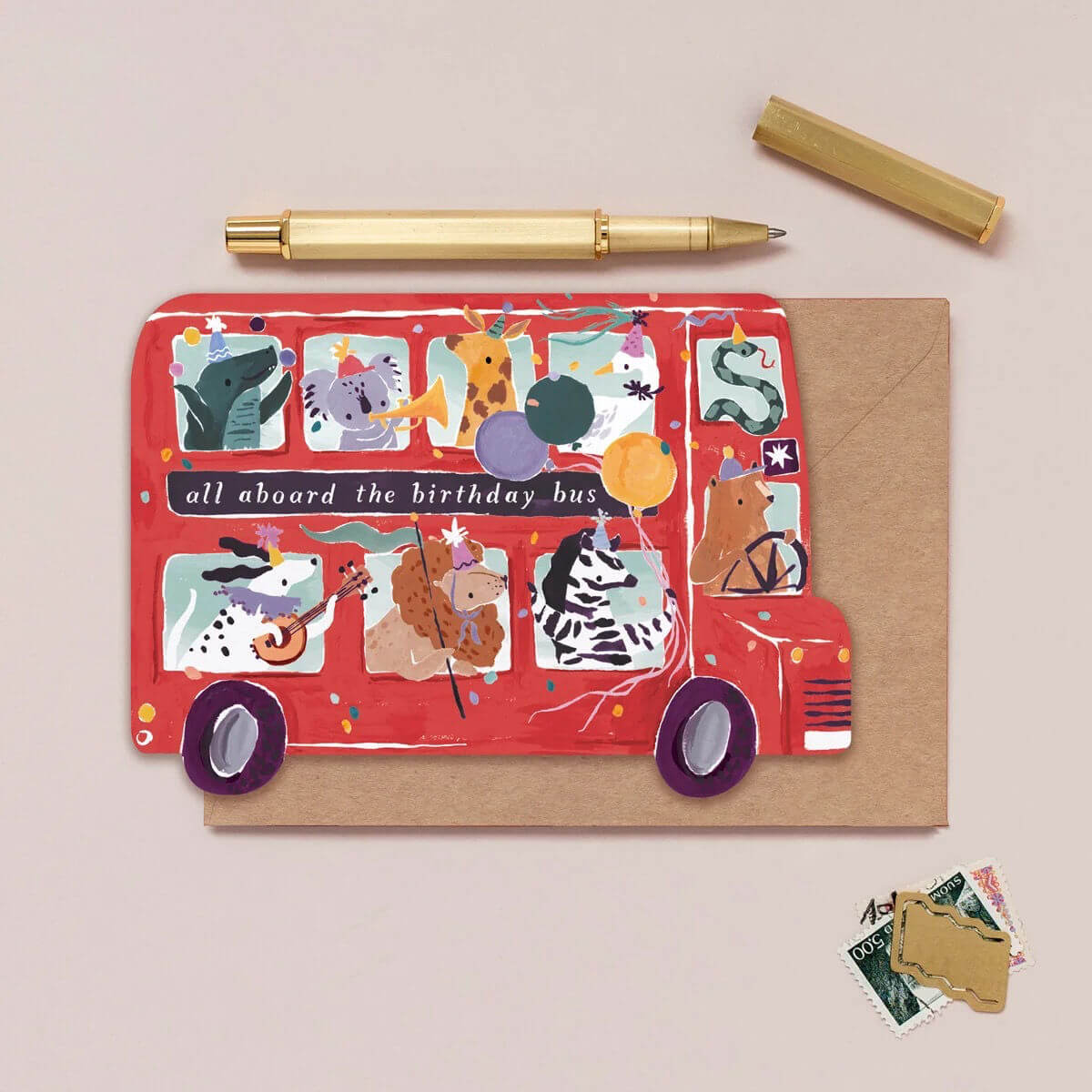 A red animal-themed birthday bus card with the text 'all aboard the birthday bus', displayed on a kraft envelope with a gold pen, gold cap, and postage stamps on a beige background.