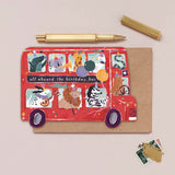 A red animal-themed birthday bus card with the text 'all aboard the birthday bus', displayed on a kraft envelope with a gold pen, gold cap, and postage stamps on a beige background.