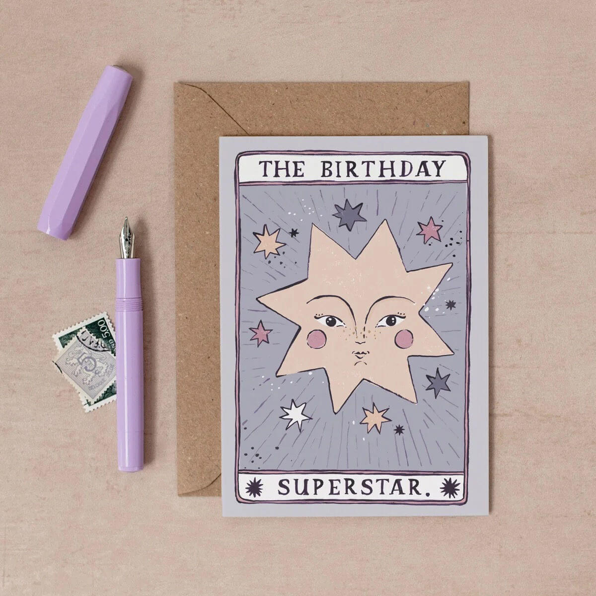 A tarot-style birthday card reading 'THE BIRTHDAY SUPERSTAR.' is displayed on a beige surface with a kraft envelope, a lavender pen, and a small postage stamp illustrated by sister paper co
