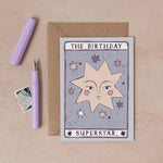 A tarot-style birthday card reading 'THE BIRTHDAY SUPERSTAR.' is displayed on a beige surface with a kraft envelope, a lavender pen, and a small postage stamp illustrated by sister paper co