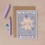 A tarot-style birthday card reading 'THE BIRTHDAY SUPERSTAR.' is displayed on a beige surface with a kraft envelope, a lavender pen, and a small postage stamp illustrated by sister paper co